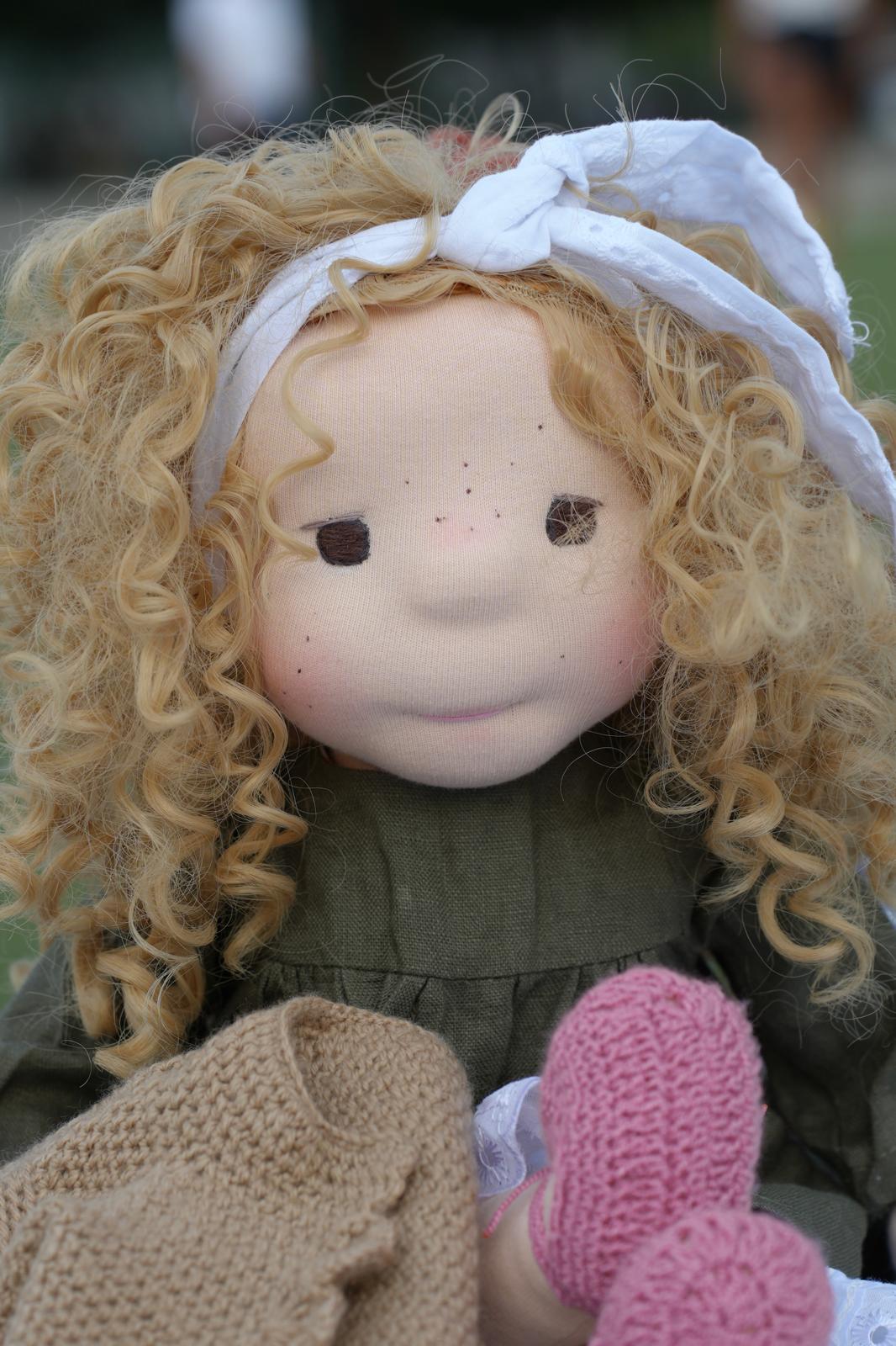 Primer plano de la muñeca de rizos rubios, con pecas y un saco de crochet en el regazo.